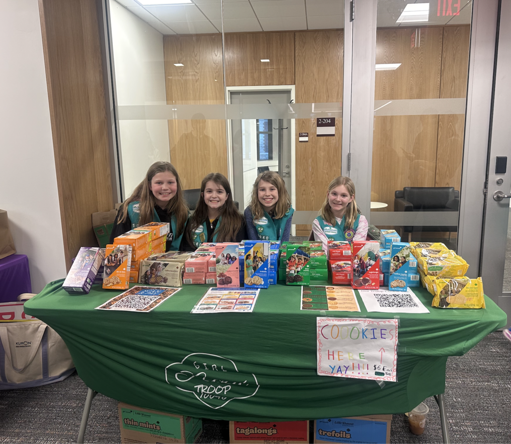 Four young girl scouts behind a green table with a variety of cookies. 